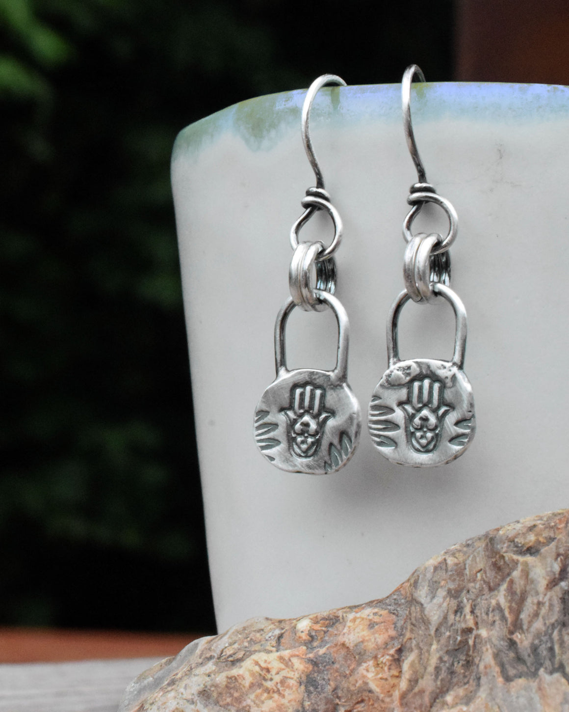 A pair of silver earrings featuring a Hamsa hand amulet design, displayed against a natural background.