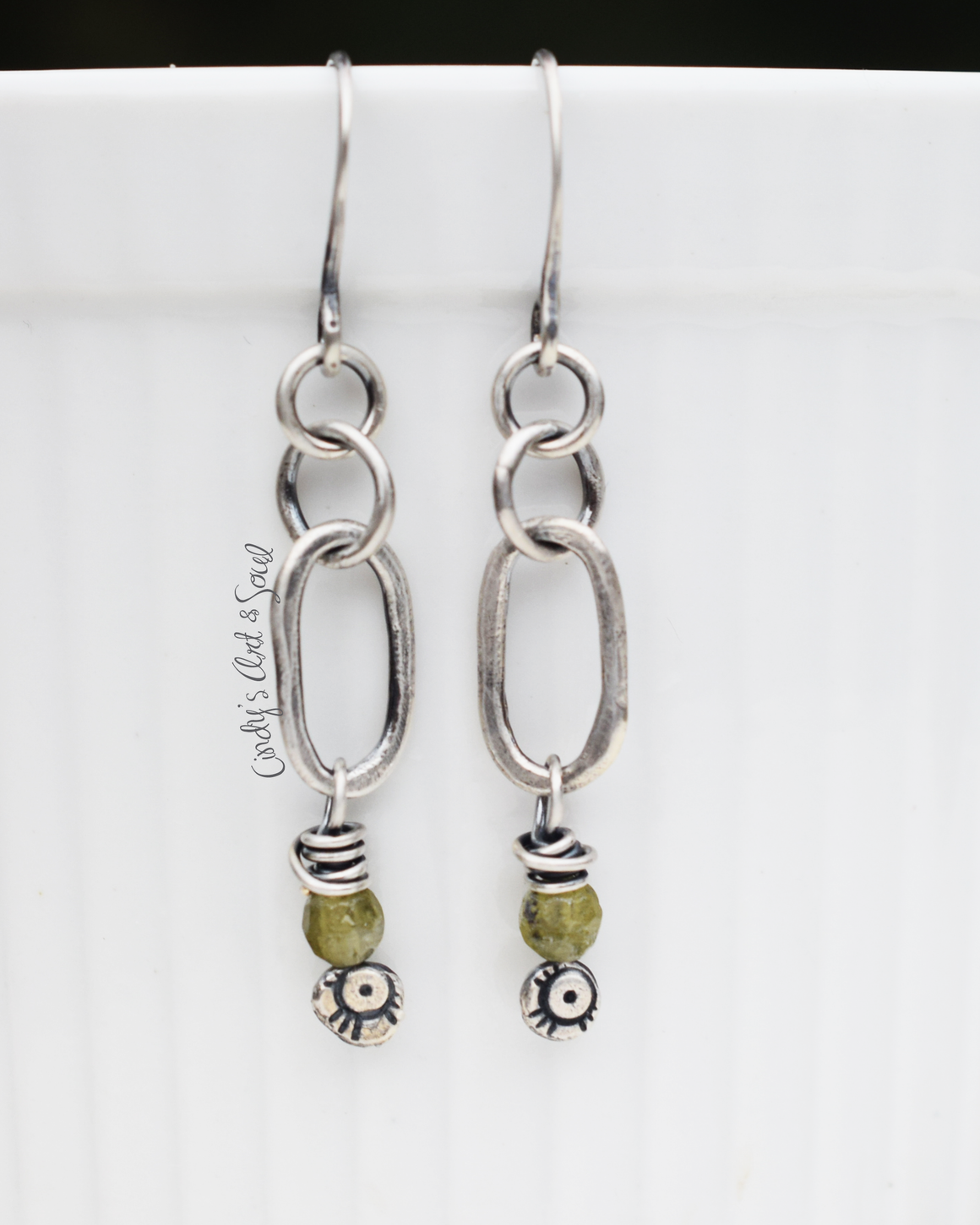 pure silver hoop earrings hanging from a white dish with dangling silver charms below with green stones on them. 