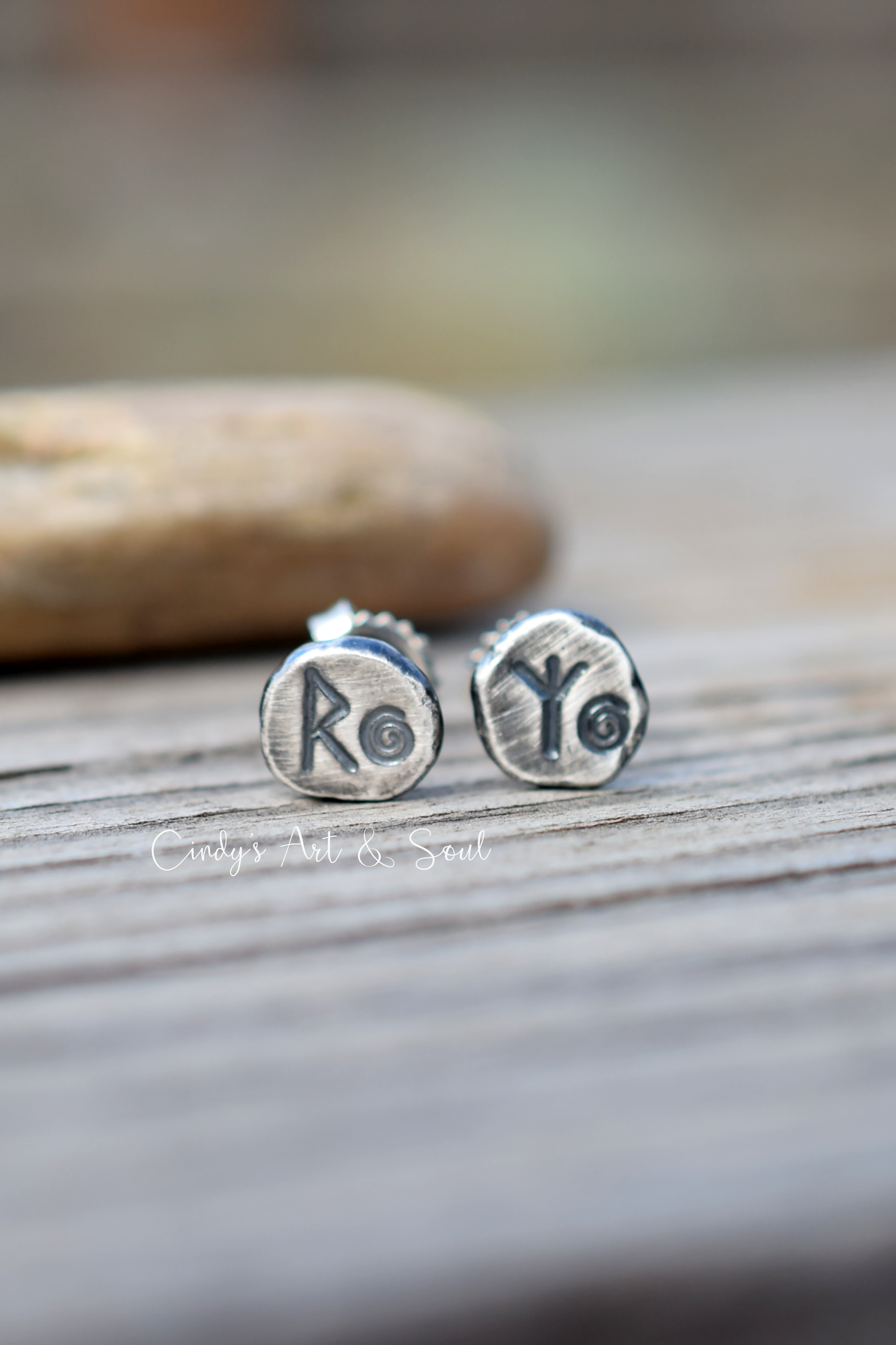 A pair of round silver stud earrings with engraved characters, resting on a weathered wooden surface with a blurred stone in the background.