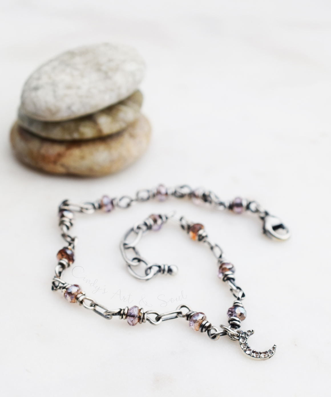 A sterling silver beaded chain bracelet with small pink glass beads and a crescent moon charm, arranged on a light surface with stacked stones in the background.