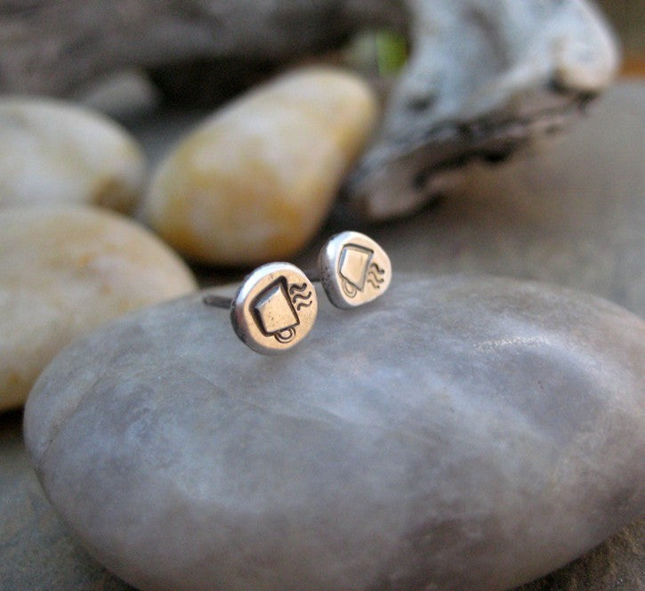 Silver post earrings with a coffee cup image on the front. The earrings are placed on a gray marbled stone. The background is more stones that are blurred. 