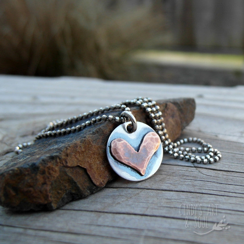 A sterling silver heart-shaped charm with a rustic finish, displayed on a wooden surface with a silver jump ring attached.