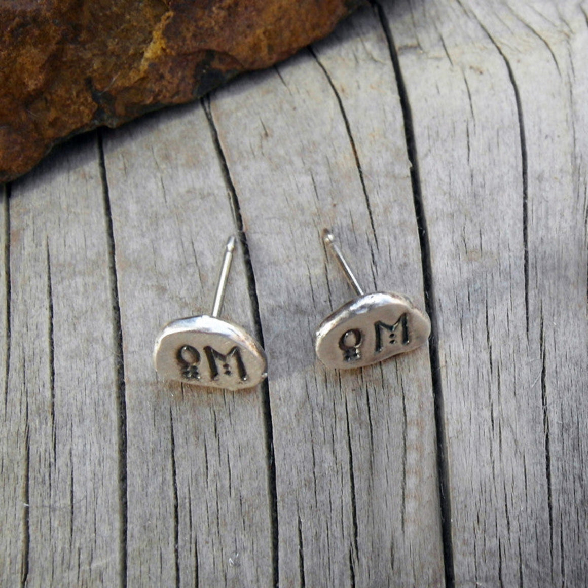 A pair of small silver stud earrings with engraved symbols, displayed on a weathered gray wooden surface with a brown rock in the corner.