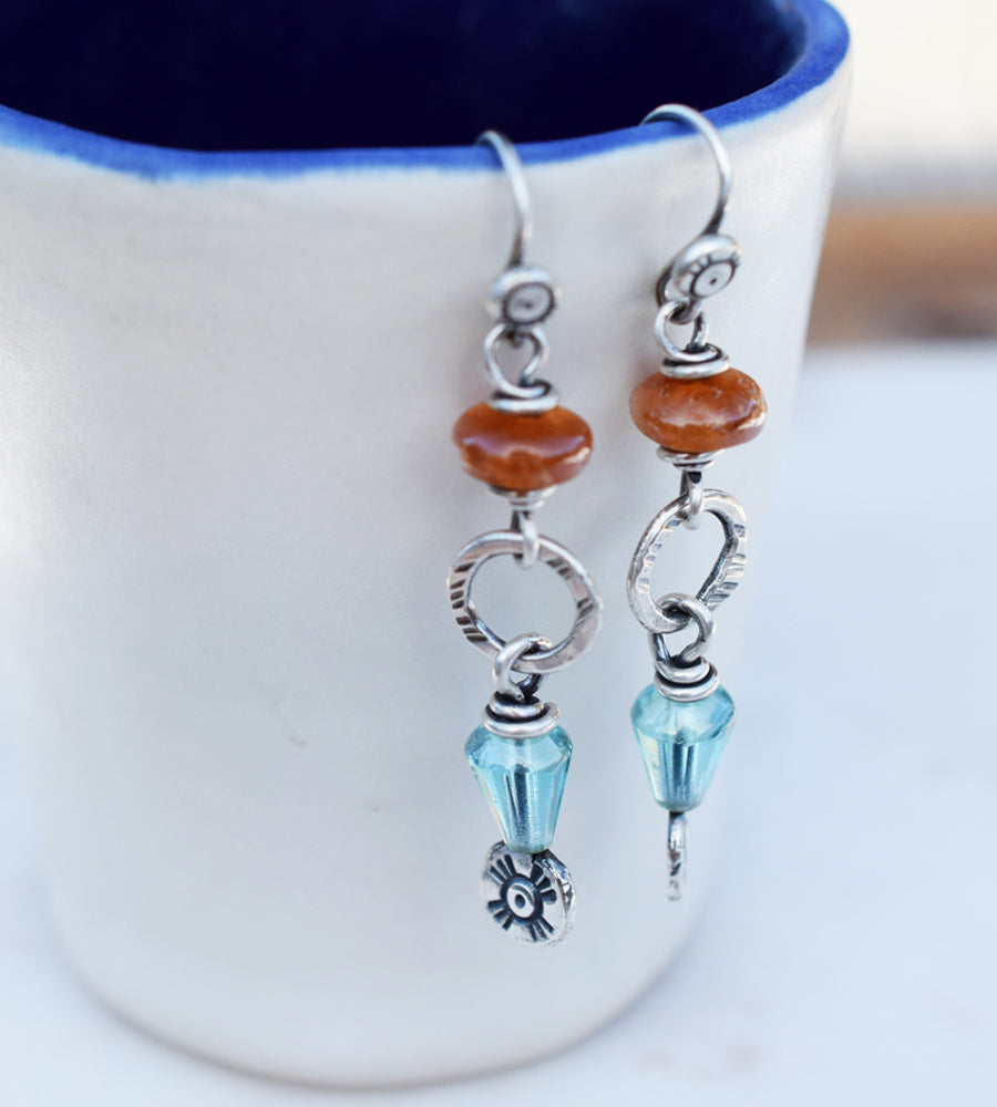 A pair of handcrafted silver boho-style dangle earrings with sun spot patterns, featuring orange and teal colors, and small silver charms.