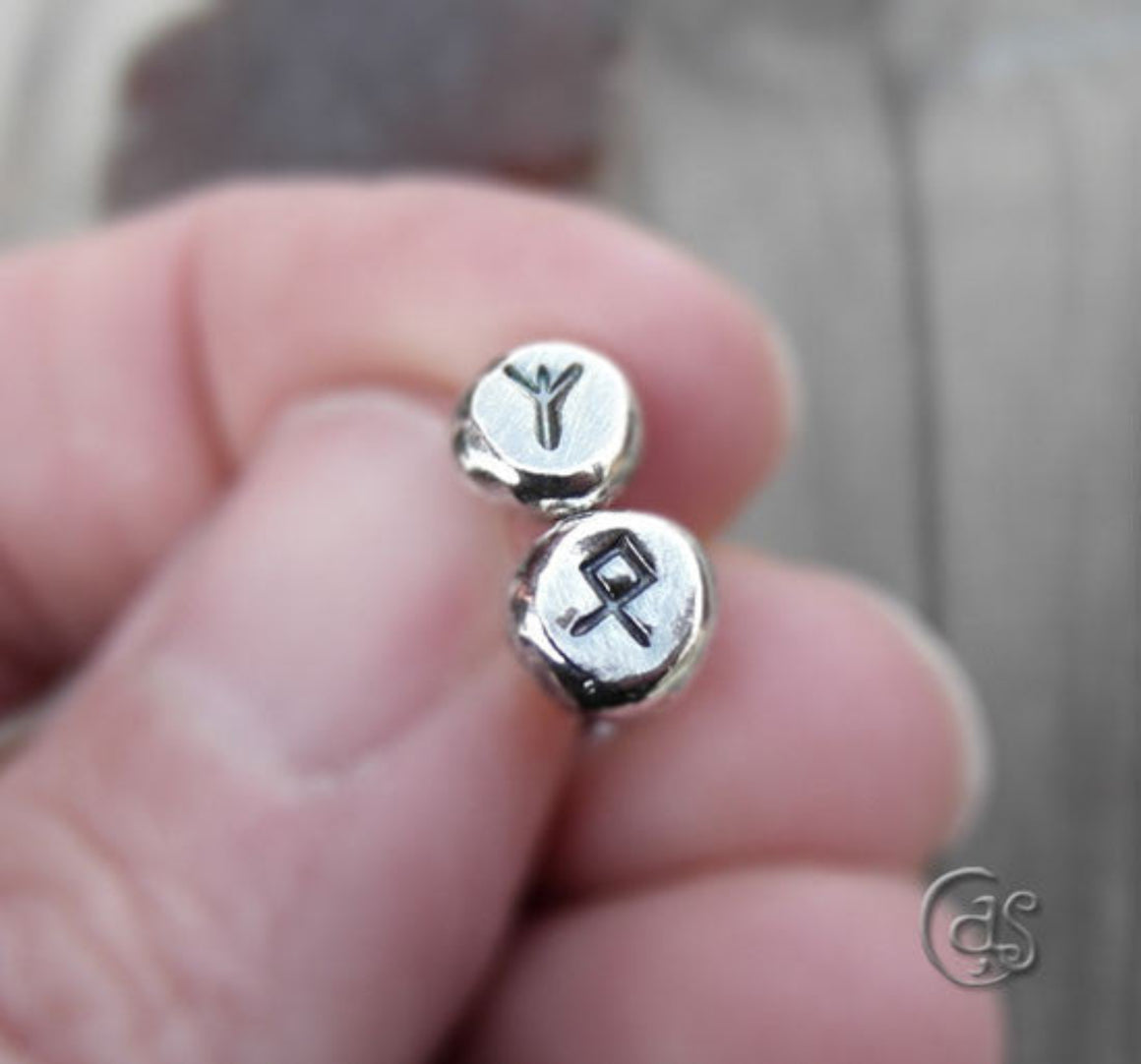 A close-up of a person holding a pair of silver rune earrings with Elder Futhark symbols inscribed on them.